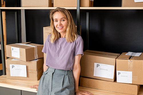 Young cheerful female worker of online shop office standing against large shelf with packed boxes with orders of clients and looking at you