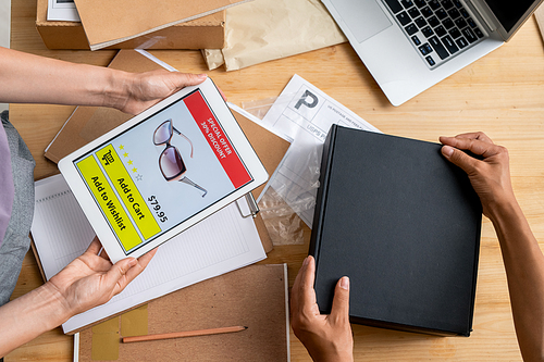 Hands of female managers of online shop holding tablet with photo of sunglasses on screen and packing black box with order of client