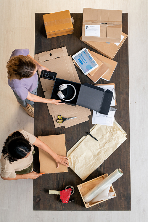 Top view of two young women standing by table and packing orders of clients among cardboards, papers and other stuff in online shop office