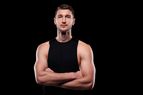 Young muscular athlete in black vest crossing his arms by chest while standing in front of camera in isolation