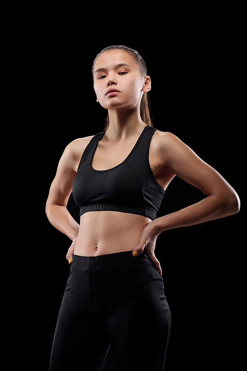 Young fit sportswoman in activewear keeping her hands on waist during physical training in isolation over black background