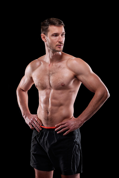 Young muscular shirtless male athlete keeping his hands on waist while standing in front of camera in isolation
