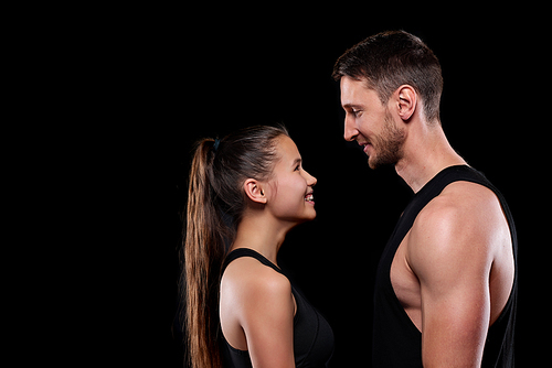 Happy young sportswoman and sportsman in activewear looking at one another with smiles over black background