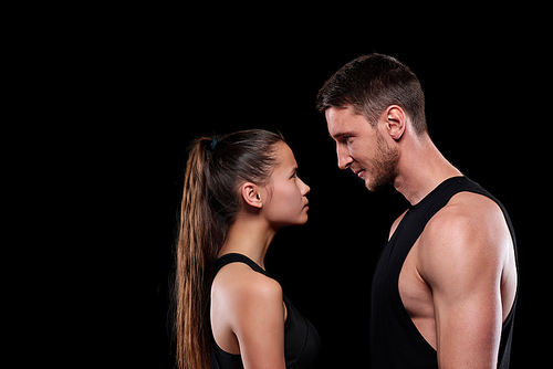 Young fit woman and man in activewear looking at one another while standing in front of camera on black background