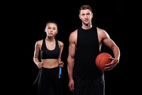 Young sporty couple in black activewear holding skipping-rope and ball while training in gym or sports center