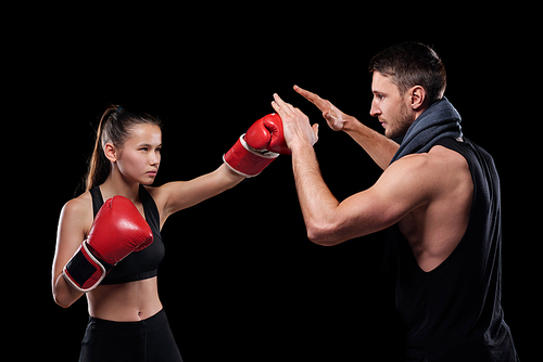 Young serious fit woman in activewear and boxing gloves hitting trainer during fight against black background
