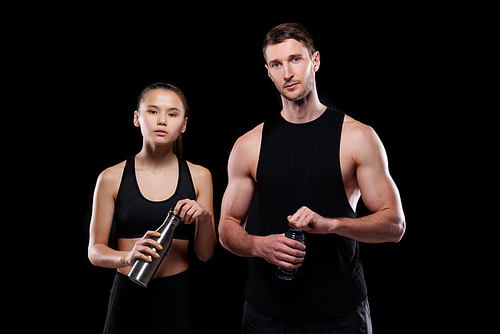 Pretty girl and muscular sportsman in activewear holding bottles with water while going to drink after training in gym