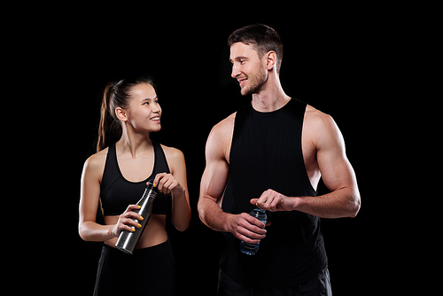 Cheerful girl and sportsman with bottles of water looking at one another with smiles while going to drink after training