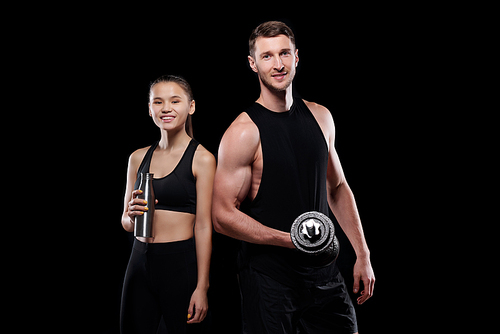 Happy young sportswoman with bottle of water and muscular man with barbell standing in front of camera