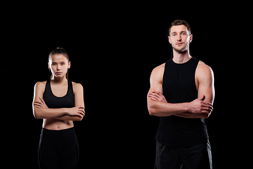 Serious young sporty couple in activewear crossing arms by chest while standing in front of camera on black background