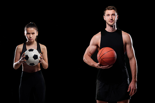 Pretty young soccer player and muscular male basketballer holding balls while standing against black background