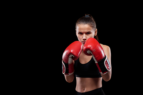 Young serious and powerful woman in activewear and boxing gloves looking at you ready to fight against black background
