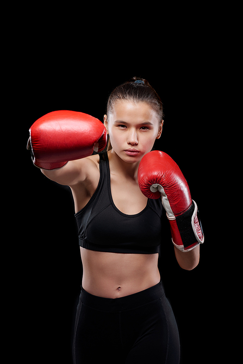 Young strong sportswoman in activewear and boxing gloves looking at you during fight against black background