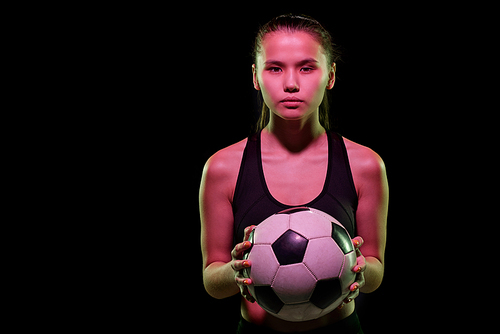 Pretty young female football player holding soccer ball while training in isolation in front of camera over black background