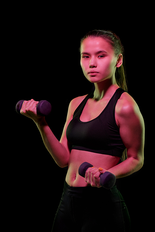 Pretty young female athlete in activewear exercising with dumbbells in front of camera over black background in isolation