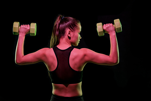 Back view of young female athlete in sportswear exercising with dumbbells in front of camera over black background