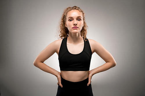 Pretty sports girl with curly blond hair keeping her hands on waist while standing in front of camera in isolation and going to work out