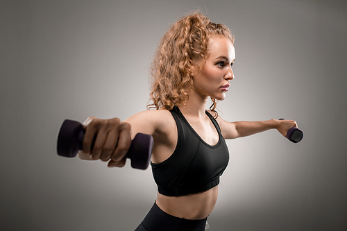 Pretty young fit female with blond curly hair outstretching her arms with dumbbells while exercising in the morning against grey background