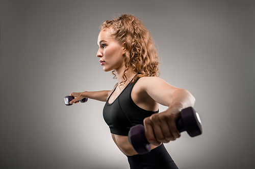 Side view of young sportswoman in black tracksuit lifting dumbbells and breathing during physical exercise against grey background