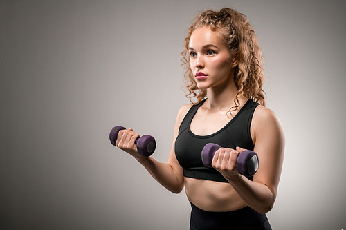 Fit girl with blond curly hair holding dumbbells with her arms bent in elbows while exercising in gym against grey background