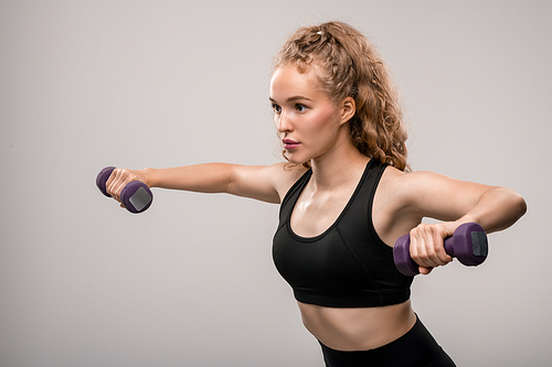 Pretty blond sportswoman in activewear standing against grey background while exercising with dumbbells during workout in isolation