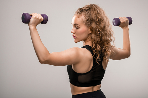 Back view of fit girl with long blond curly hair exercising with dumbbells with her arms bent in elbows against grey background in isolation