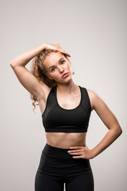 Fit young woman in black tracksuit touching her long blond curly hair and keeping another hand on waist against grey background in isolation