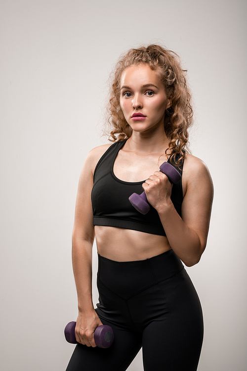 Pretty girl with blond curly hair holding dumbbells in hands while doing exercise for arm muscles during workout in isolation