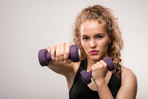 Pretty young sportswoman with blond curly hair stretching arm with dumbbell while exercising in the morning against grey background