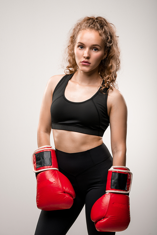 Blond fit girl in black tracksuit and red boxing gloves standing in front of camera in isolation ready for training in sports club or gym