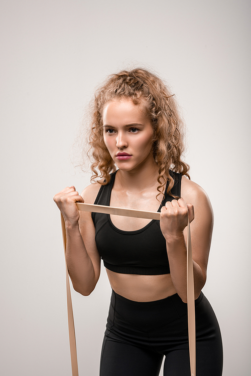 Young sportswoman with blond curly hair stretching elastic band while exercising in gym against grey background during training