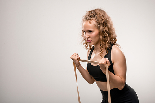 Young sportswoman with blond curly hair making effort while exercising with elastic band against grey background during training