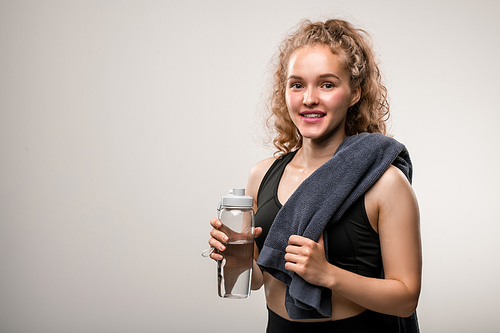 Happy fit woman in black tracksuit holding bottle of water and soft towel while having rest after sports training against grey background