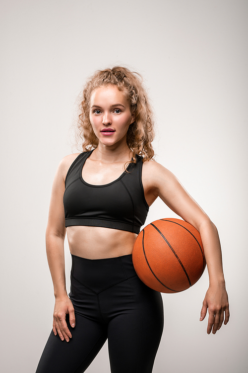 Young blond active female in sportswear holding ball between her waist and arm while standing in front of camera on grey background