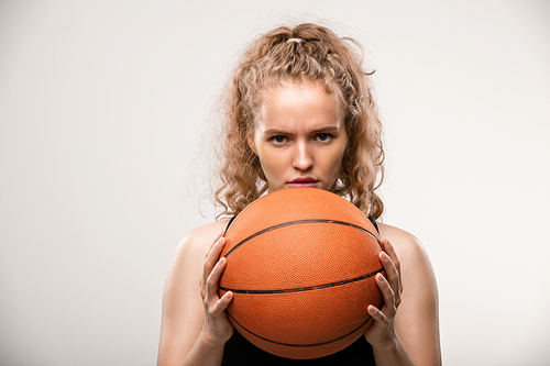Young female basketball player holding ball by her face while standing in front of camera and looking at rival over grey background