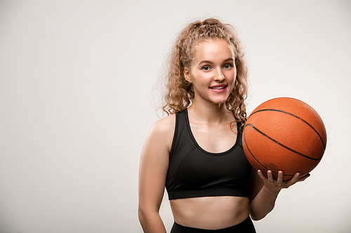 Happy young successful female basketball player in black tracksuit holding ball in left hand while standing in front of camera in isolation