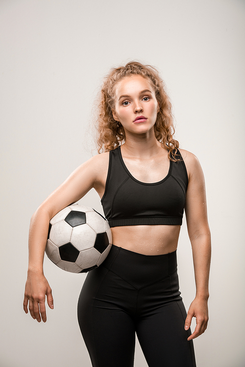 Young active sportswoman in tracksuit holding soccer ball between her waist and arm while standing in front of camera on grey background