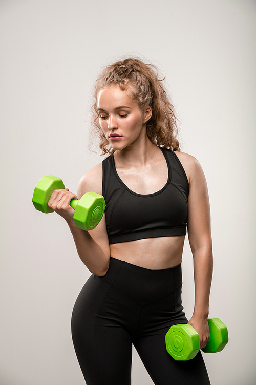 Fit girl in black sportswear holding green dumbbells in hands while bending one arm during physical exercise on grey background