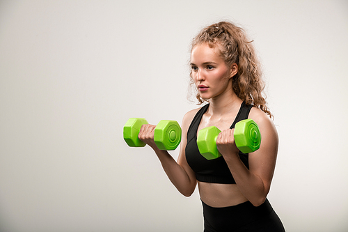Pretty blond fit girl with curly hair doing exercise for arm muscles with dumbbells during workout in front of camera in isolation