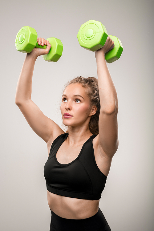 Active blond girl in black sportswear holding green dumbbells in hands over her head during physical exercise against grey background