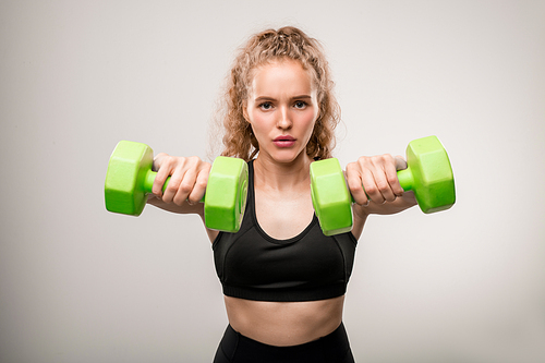 Young active sportswoman in black tracksuit standing in front of camera and looking at you while doing exercise with green dumbbells