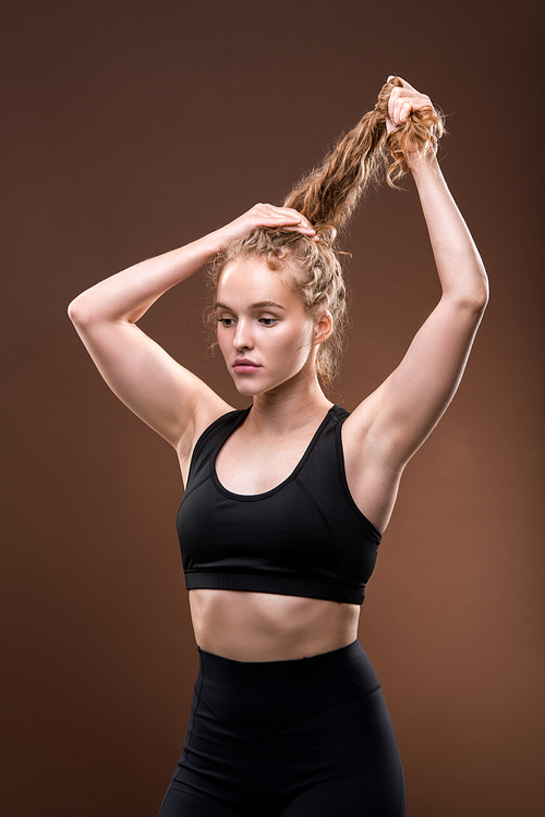 Slim young woman in black tracksuit holding her long blond curly hair while making ponytail before workout against brown background