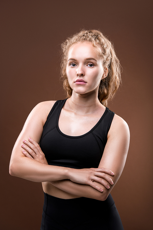 Slim young serious sportswoman in black tracksuit keeping her arms crossed by chest while standing in front of camera in isolation