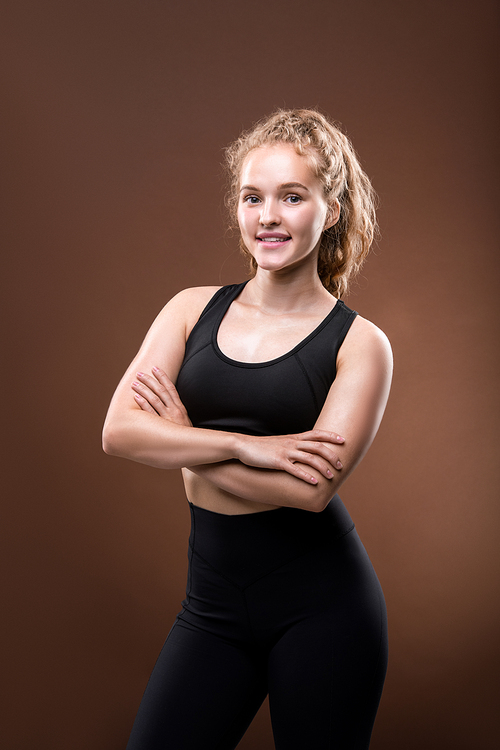 Young cheerful blond sportswoman in black tracksuit keeping her arms crossed by chest and looking at you with smile on brown background