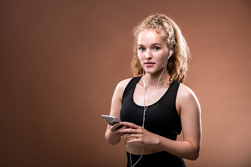 Young blond sportswoman in activewear and earphones scrolling in smartphone while choosing track from playlist before workout