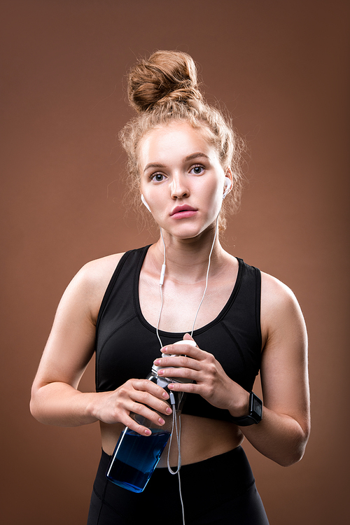 Young fit woman in earphones and black tracksuit holding bottle of water while having rest after sports training against brown background
