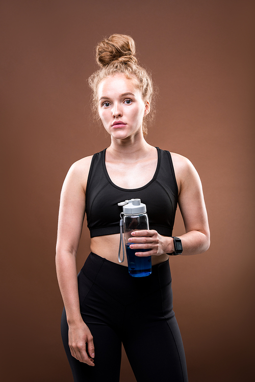 Pretty sportswoman with blond curly hair holding plastic bottle with blue drink while having break between workouts in isolation