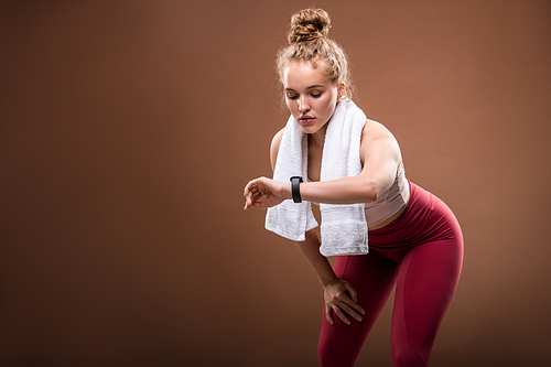 Tired young blond sportswoman in activewear looking at fitbit on her wrist while doing difficult physical exercise against brown background