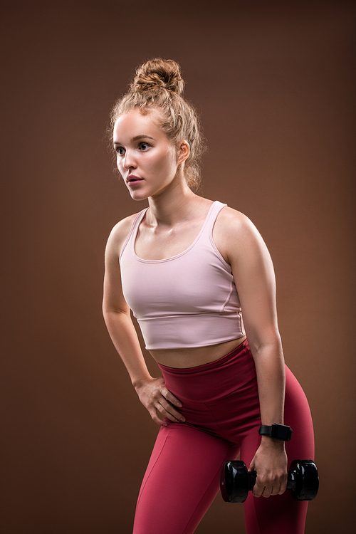 Active blond girl in sportswear holding dumbbell in left hand with the right one on hip during physical exercise against brown background