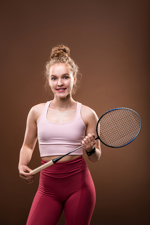 Young cheerful blond female tennis player in pink tanktop and crimson leggins holding racket while standing in front of camera in isolation